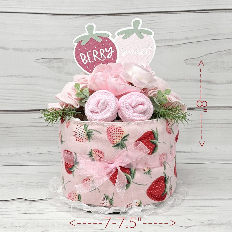 May include: A pink and white diaper cake decorated with pink flowers, green leaves, and two strawberry-shaped signs that say "Berry sweet". The cake is 7.5 inches in diameter and 8 inches tall.