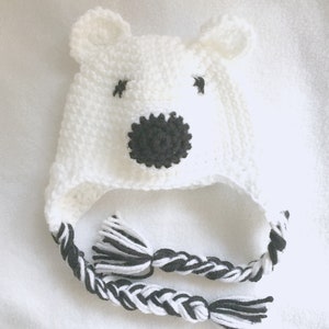 Peut inclure: Chapeau en crochet blanc en forme d'ours polaire avec des yeux noirs, un nez noir et des liens en laine noire et blanche tress&eacute;s.