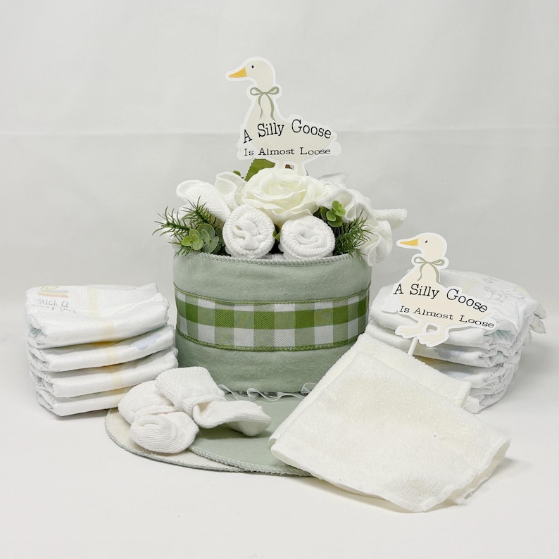 May include: A baby gift set featuring a green and white gingham-patterned container with a floral arrangement and the text "A Silly Goose Is Almost Loose." The set includes diapers, washcloths, and baby socks.