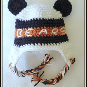 Peut inclure: Bonnet b&eacute;b&eacute; en crochet blanc avec des rayures noires et orange et des pompons noirs. Le bonnet est brod&eacute; du mot "BEARS" en orange sur une bande noire.