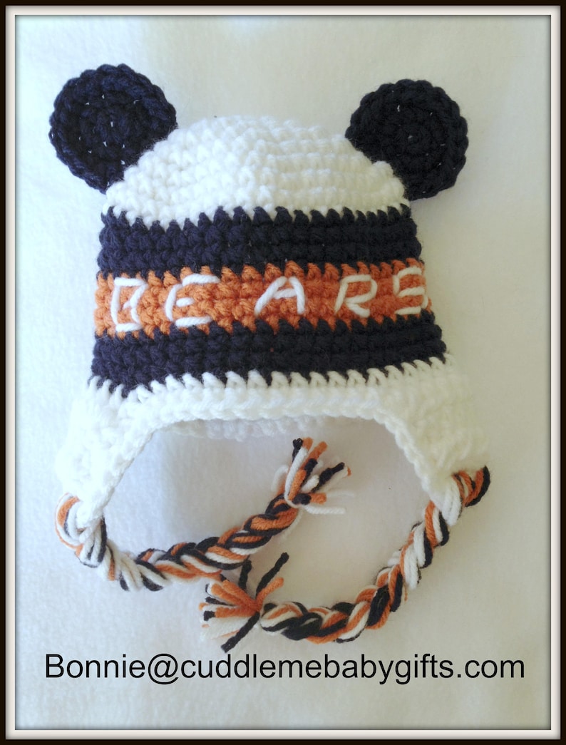 Peut inclure: Bonnet b&eacute;b&eacute; en crochet blanc avec des rayures noires et orange et des pompons noirs. Le bonnet est brod&eacute; du mot "BEARS" en orange sur une bande noire.