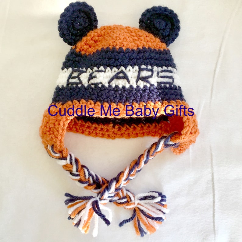 May include: A crocheted baby hat with ear flaps and a long braided tie. The hat is orange, navy blue, and white with the word "BEARS" crocheted on the front.