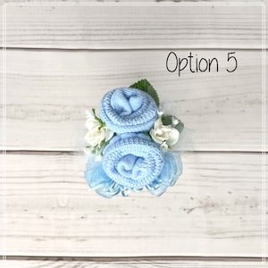 May include: Two light blue baby socks rolled into the shape of roses, with white flowers and blue ribbon, on a white wooden background. Option 5.