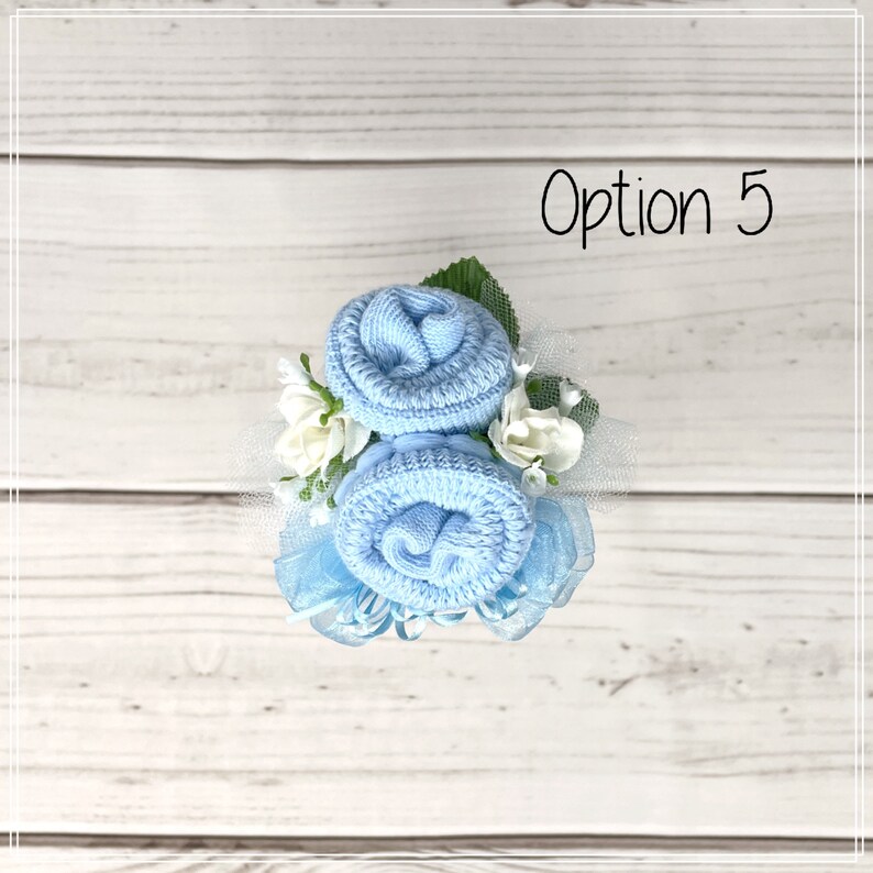 May include: Two light blue baby socks rolled into the shape of roses, with white flowers and blue ribbon, on a white wooden background. Option 5.
