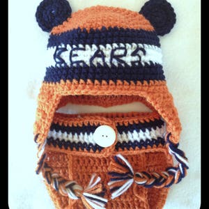 Peut inclure: Un bonnet b&eacute;b&eacute; en crochet avec des rabats d'oreilles et un bouton blanc. Le bonnet est orange, blanc et bleu marine avec le mot "Bears" en lettres blanches sur le devant.