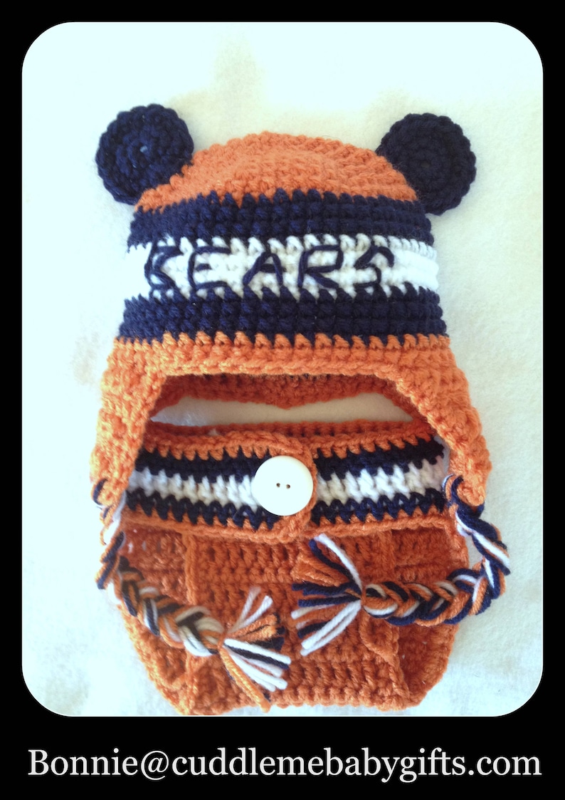 Peut inclure: Un bonnet b&eacute;b&eacute; en crochet avec des rabats d'oreilles et un bouton blanc. Le bonnet est orange, blanc et bleu marine avec le mot "Bears" en lettres blanches sur le devant.