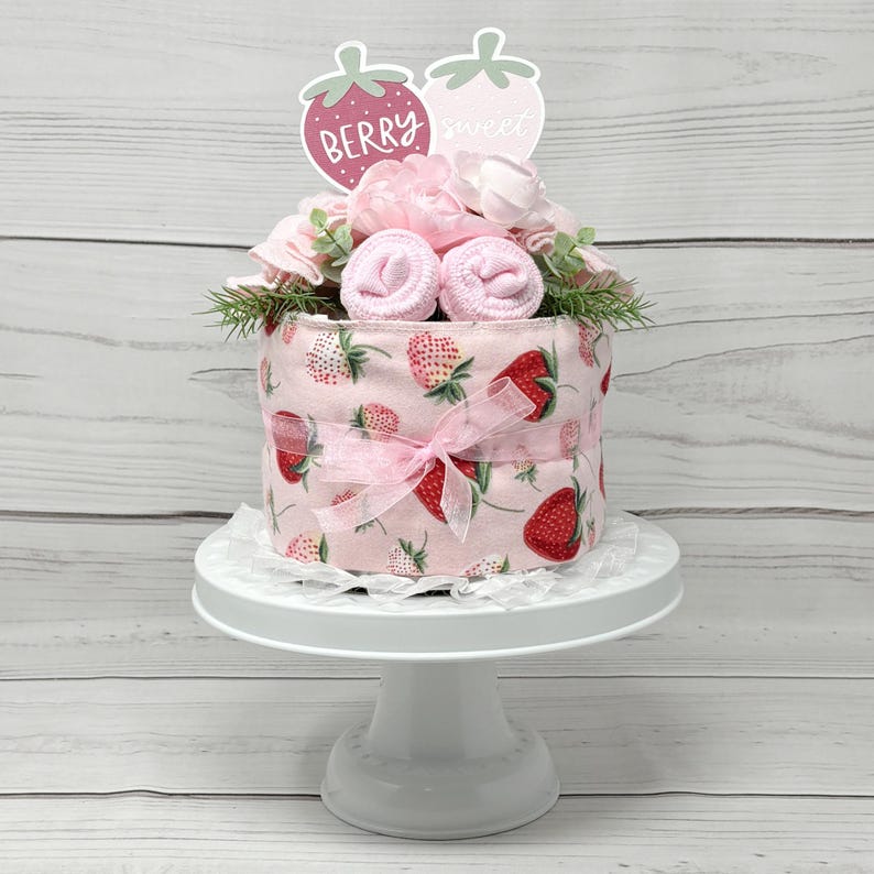 May include: A pink and white diaper cake decorated with strawberries and a topper that says "Berry Sweet".