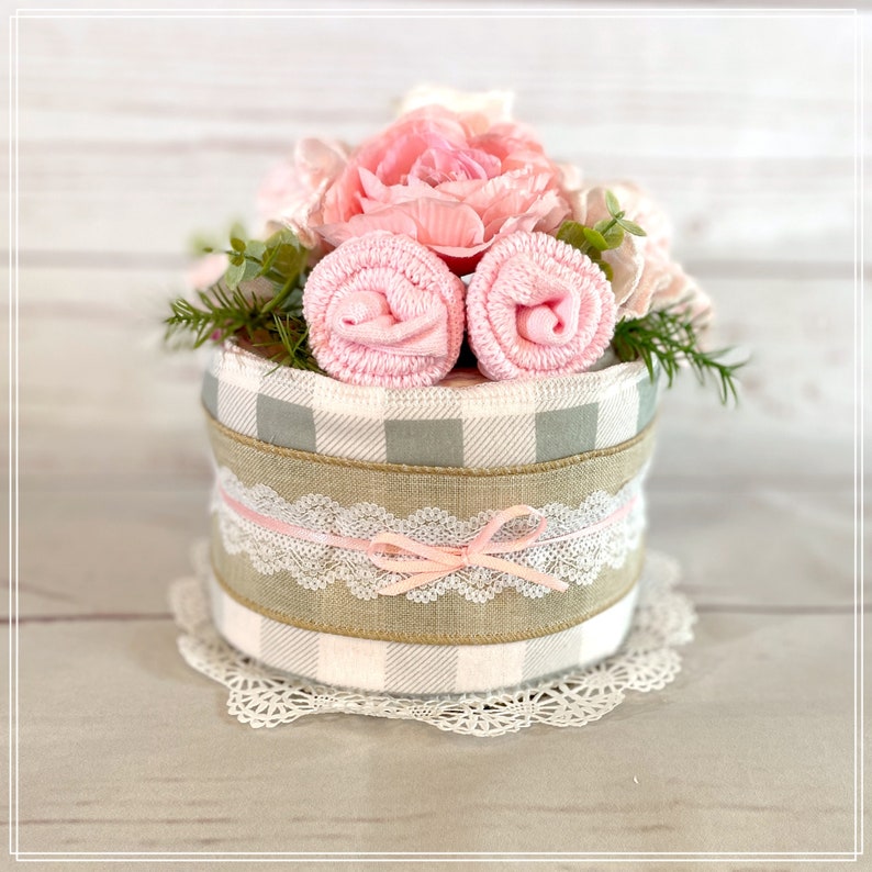 May include: A baby shower gift wrapped in a gray and white checkered pattern with a burlap ribbon and a pink lace ribbon. The gift is topped with pink flowers and two rolled up pink towels.