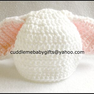 Peut inclure: Chapeau de b&eacute;b&eacute; en crochet blanc avec des oreilles en crochet rose. Le chapeau a un motif de point textur&eacute;. Le texte "cuddlemebabygifts@yahoo.com" est visible sur le chapeau.