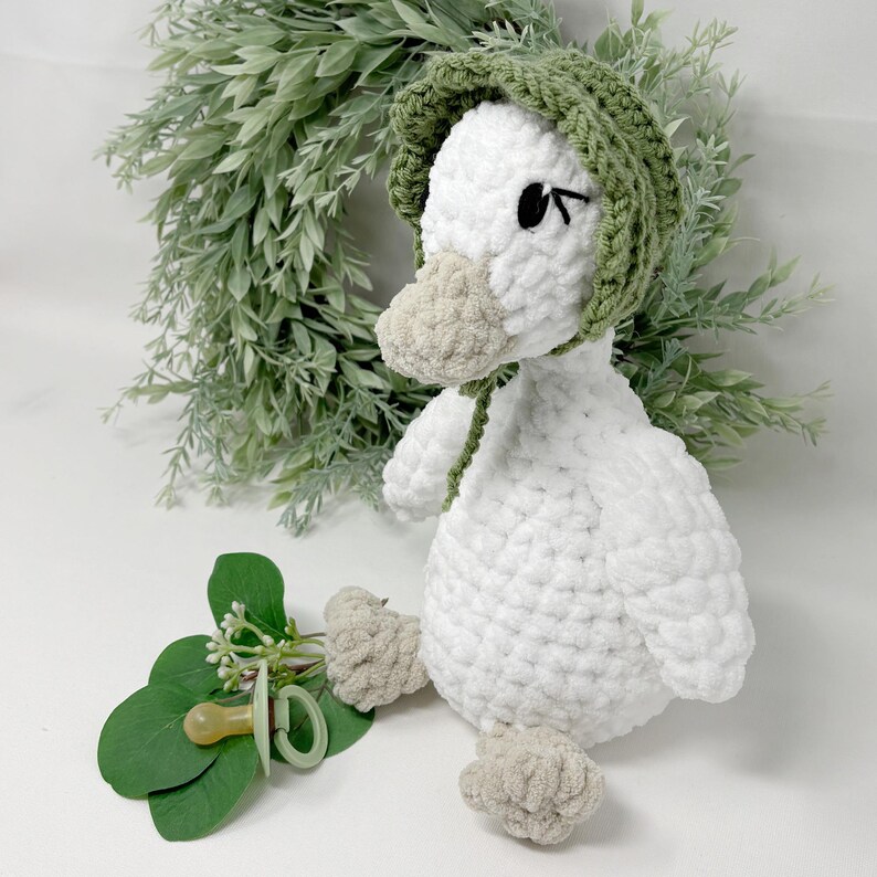 May include: A handmade white crochet duck wearing a green bonnet. The duck has a beige beak and feet, with black eyes. A pacifier and greenery are in the foreground. The duck is a soft toy.