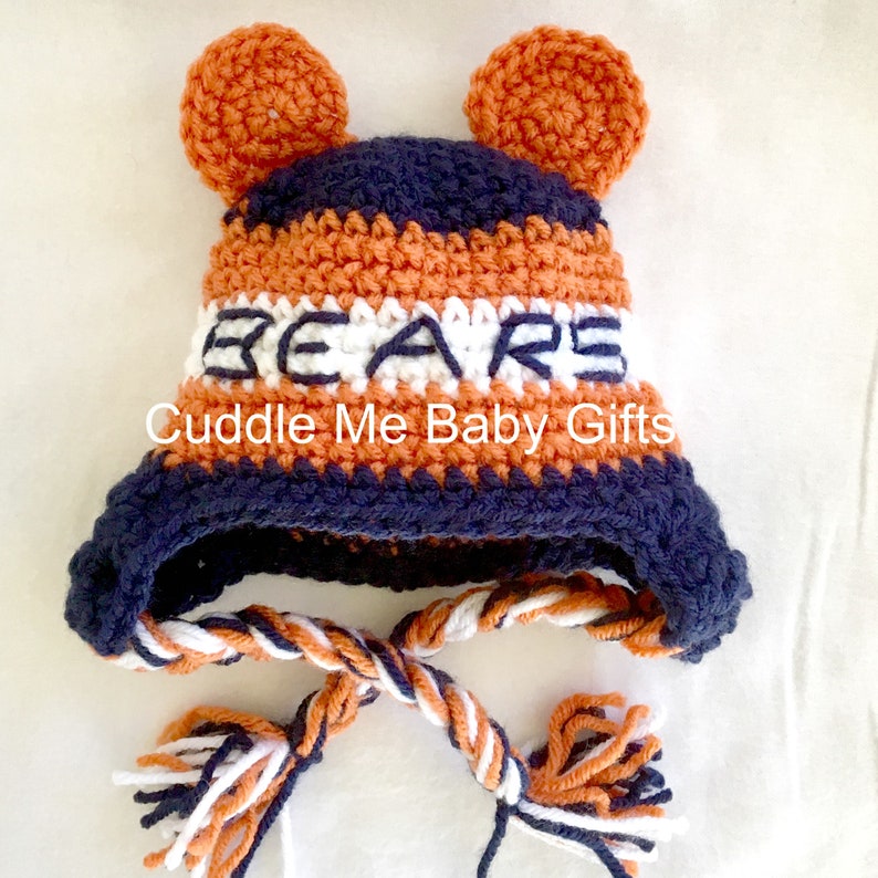 May include: A crocheted baby hat with orange, white, and navy blue stripes. The hat has two orange ear flaps and a white stripe with the word "BEARS" embroidered in navy blue. The hat has a long navy blue, white, and orange braided tie.