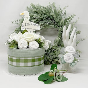 May include: A baby shower centerpiece featuring a light green felt cylinder with a green and white gingham ribbon. It is decorated with white roses, rolled washcloths, and a sign that reads "A Silly Goose Is Almost Loose."