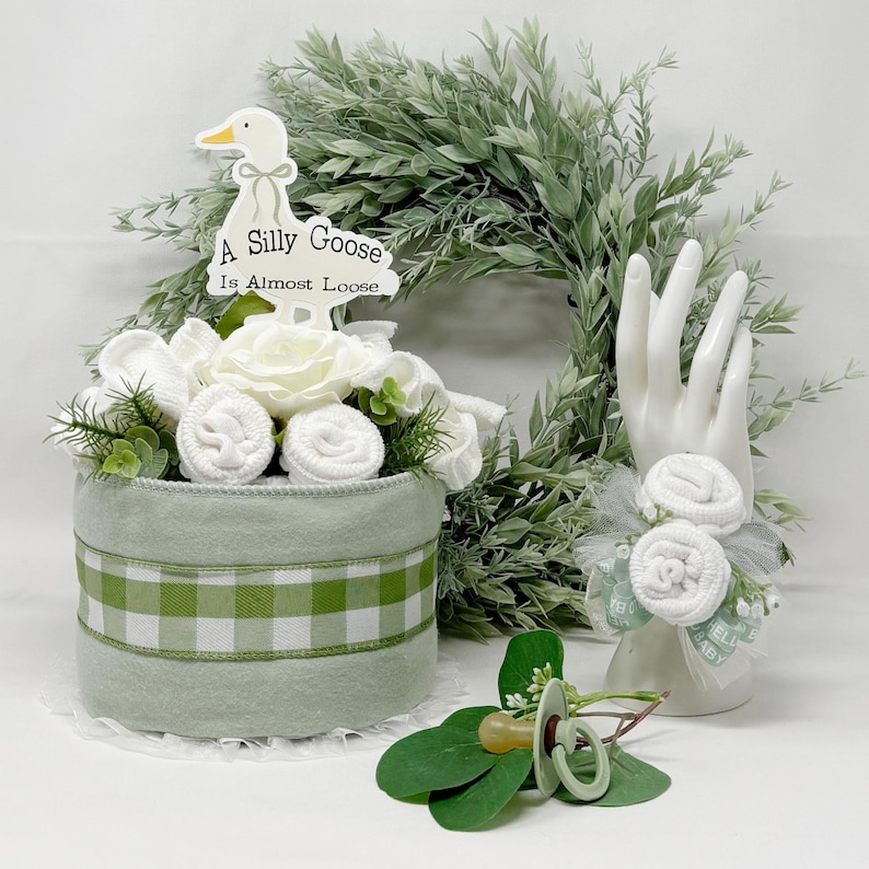 May include: A baby shower centerpiece featuring a light green felt cylinder with a green and white gingham ribbon. It is decorated with white roses, rolled washcloths, and a sign that reads "A Silly Goose Is Almost Loose."