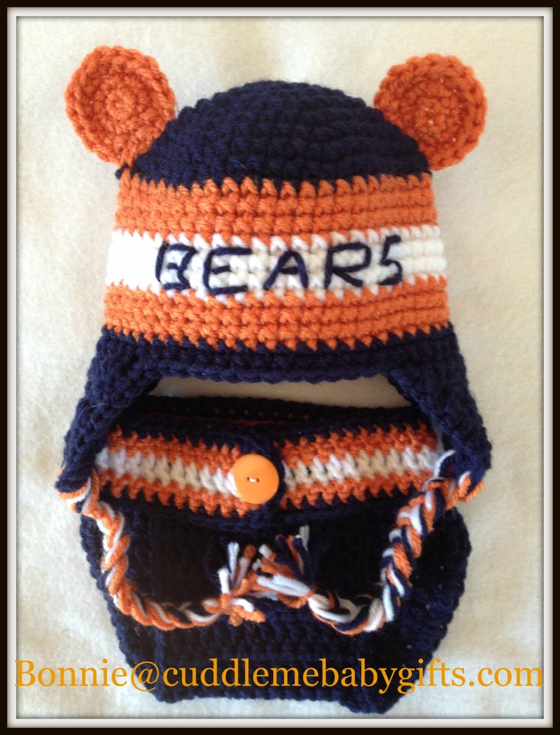 Peut inclure: Un ensemble de bonnet et de couverture de couche pour b&eacute;b&eacute; en crochet bleu marine, orange et blanc avec le mot "BEARS" en blanc sur le bonnet. Le bonnet a deux oreilles orange.