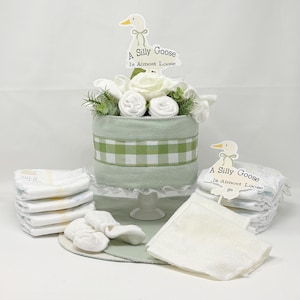 May include: A baby shower gift set featuring a light green cake-shaped centerpiece with white flowers and rolled washcloths. The cake has a green and white checkered ribbon. Diapers, socks, and a burp cloth surround the cake. The sign says "A Silly Goose Is Almost Loose."