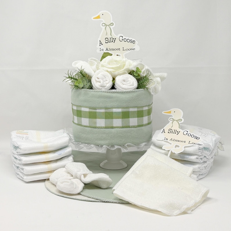 May include: A baby shower gift set featuring a light green cake-shaped centerpiece with white flowers and rolled washcloths. The cake has a green and white checkered ribbon. Diapers, socks, and a burp cloth surround the cake. The sign says "A Silly Goose Is Almost Loose."