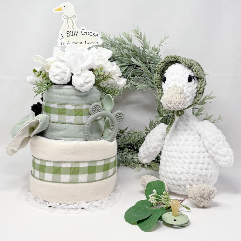 May include: A baby shower gift set featuring a diaper cake with green gingham accents, a crocheted goose, and a pacifier. The diaper cake has a topper that reads "A Silly Goose Is Almost Loose". The set includes a green teething ring and a white floral arrangement.