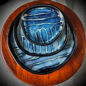 May include: A set of three nested ceramic plates with a glossy finish, featuring a deep blue and black glaze with white, wave-like linear patterns. Each plate has a distinct black rim and a slightly irregular, organic circular shape.