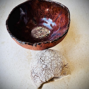 May include: A handmade ceramic bowl with a deep red interior and a textured, cracked white ceramic piece. The bowl has a dark rim and a unique, organic shape. The ceramic piece has a cracked pattern.