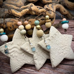 May include: Cream-colored star-shaped ornaments with embossed floral patterns, strung on brown twine. Each star has a unique beaded accent in various colors, including gold, turquoise, and white. The ornaments are displayed against a wooden background.