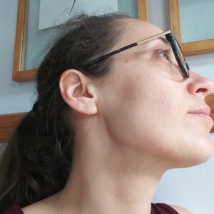 May include: A close-up of a person's ear with two small gold stud earrings. The person is wearing glasses and has dark hair.
