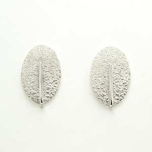 Peut inclure: Une paire de boucles d'oreilles ovales en argent avec une surface texturée et une ligne verticale au centre.