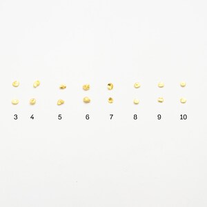 May include: Twelve rows of small, round, gold-colored beads arranged in a line on a white background. The rows are numbered 1 through 12.