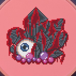May include: Cross-stitch embroidery on a pink hoop featuring a stylized eye with red, gray, and black details. The design is surrounded by embroidery floss in purple, blue, and red, along with a pair of decorative scissors.