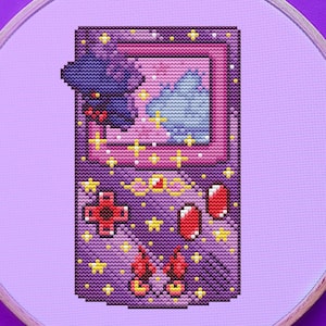 Puede incluir: Un aro de bordado de punto de cruz morado y rosa con una imagen pixelada de una consola de videojuegos portátil. La consola tiene un fondo morado y rosa con estrellas amarillas y un cartucho de juego rojo y amarillo.