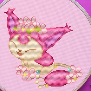 May include: A completed cross-stitch project featuring a pink cartoon cat with a flower crown, set in a wooden embroidery hoop. Pink embroidery floss and ornate scissors are arranged around the hoop on a magenta surface. Craft supplies.