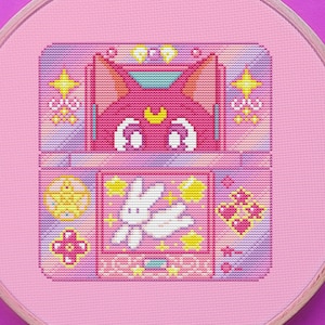 May include: A pink and white cross-stitch embroidery hoop featuring a pixelated design of a pink cat with a crescent moon on its forehead, a white rabbit with wings, and a pink heart. The design is surrounded by stars and hearts.