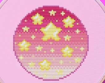 May include: A cross-stitch embroidery hoop with a pink background. The design features a circle with yellow stars on a gradient pink background. Pink embroidery floss and scissors are also visible.