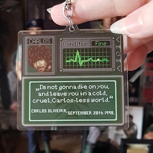 May include: A green and black pixelated keychain featuring a character named Carlos. The keychain includes a quote from the character: "I'm not gonna die on you, and leave you in a cold, cruel, Carlos-less world." The quote is attributed to Carlos Oliveira, September 28th 1998.