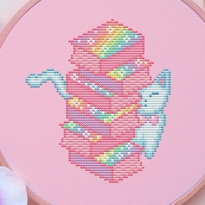 May include: A cross-stitch embroidery hoop featuring a colorful, pixelated design of a teapot and a bunny. The design incorporates pink, blue, yellow, and rainbow colors. The background is pink, and the hoop is surrounded by a black dish, chopsticks, and flowers.