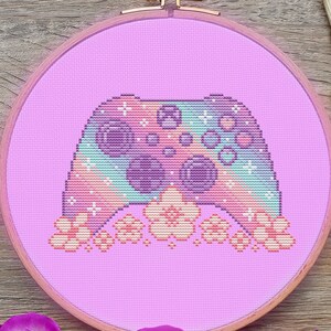 May include: A pink and purple cross-stitch embroidery hoop with a colorful pixelated video game controller design. The controller is surrounded by small pink flowers.