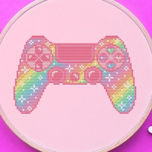 May include: A cross-stitch embroidery hoop with a pink video game controller design and rainbow accents. The design is set against a pink fabric background. Pink embroidery thread and silver scissors are also visible.