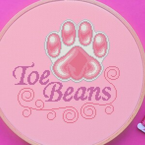 May include: A pink cross-stitch embroidery hoop with a paw print design and the words "Toe Beans". The image includes pink embroidery floss and decorative scissors, all set against a bright pink background.