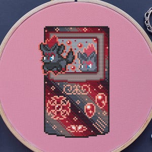 Puede incluir: Bordado de punto de cruz de una consola de juegos portátil retro con un diseño de criatura pixelada. El diseño está dentro de un bastidor de bordado rosa, con hilo de bordar y tijeras cerca. La paleta de colores incluye tonos de rojo, negro y rosa.