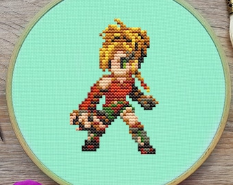 May include: Cross-stitch embroidery hoop with a pixelated image of a female character with blonde hair and a red and green outfit. The hoop is on a wooden surface with pink flowers.