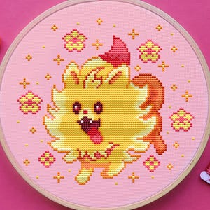 以下が含まれることがあります： ピンク色の背景に、黄色の漫画の犬が描かれたクロスステッチ刺繍枠。ピンクとオレンジの色合いの刺繍糸、銀色のハサミが写っています。犬は舌を出しており、星と花の模様に囲まれています。