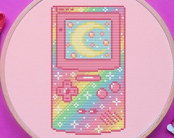 May include: A cross-stitch embroidery hoop featuring a pink retro handheld game console design with a moon and stars. Pink embroidery floss and silver scissors are arranged around the hoop on a magenta surface.
