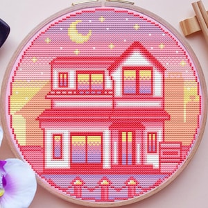 Peut inclure: Un cercle de broderie au point de croix représentant une maison rose sous un ciel étoilé et un croissant de lune. L'œuvre est dans un cercle en bois, avec une petite assiette noire et des baguettes à proximité. Des orchidées aux pétales violets et blancs sont également présentes.