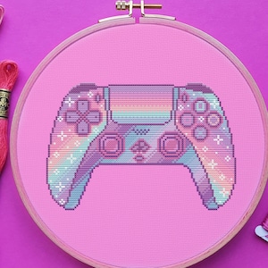 May include: A pink and white cross-stitch embroidery hoop featuring a rainbow-coloured video game controller design. The hoop is surrounded by pink embroidery floss and a pair of silver embroidery scissors.