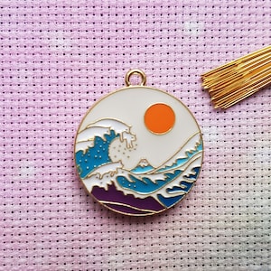May include: A white enamel charm with a gold rim depicting a blue and white wave with a purple mountain and an orange sun. The charm is on a pink and white textured background.