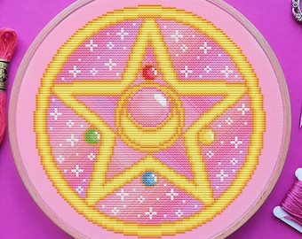 May include: A pink and yellow cross-stitch embroidery of a five-pointed star with a crescent moon and a large gem in the center. The embroidery is framed in a wooden hoop and surrounded by pink embroidery floss.