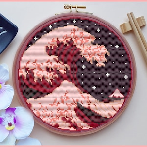 May include: A red and white cross-stitch embroidery of the Great Wave off Kanagawa, a famous Japanese woodblock print by Hokusai, framed in a light pink wooden hoop.