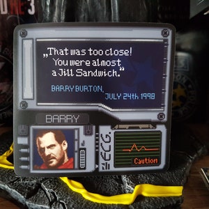 K&ouml;nnte beinhalten: Ein schwarzer und grauer Videospiel-Aufkleber mit einem pixeligen Bild eines Mannes mit einem roten Hemd vor einem schwarzen Hintergrund. Der Text auf dem Aufkleber lautet "That was too close! You were almost a Jill Sandwich." Barry Burton, July 24th 1998. Der Aufkleber zeigt auch ein pixeliges Bild eines Herzfrequenzmonitors mit dem Wort "Caution" darunter.