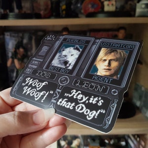 May include: A black and white sticker with a pixelated design. The sticker features two images, one of a white wolf and the other of a man. The text on the sticker reads "PUEBLOS", "USSTRATCOM", "BEST BOY", "AGENT", "DOG [LEON]", "Woof", "Woof!" and "Hey, it's that Dog!"