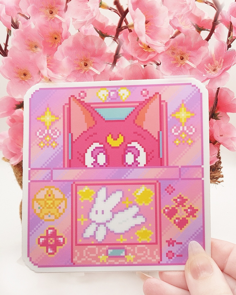 May include: A pink pixel art sticker featuring a pink cat with a crescent moon on its forehead, a white bunny with wings, and a pink heart design. The sticker is surrounded by a pink and purple border with stars and hearts.