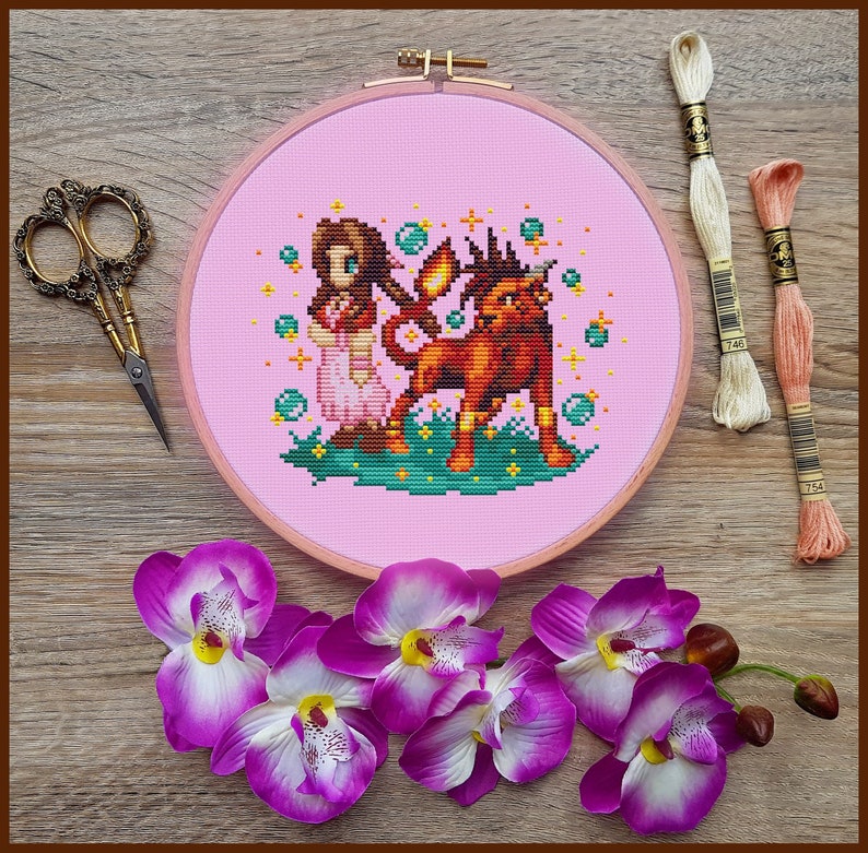 Puede incluir: Un aro de bordado de punto de cruz rosa con un dise&ntilde;o de una ni&ntilde;a con el pelo largo y casta&ntilde;o y un vestido rojo de pie junto a un le&oacute;n rojo con una melena amarilla. El fondo es azul claro con estrellas blancas y burbujas.